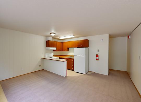 A spacious living area here at Country Lane showcasing soft carpet flooring, neutral walls, and an open layout that flows into the kitchen, which features warm wood cabinetry, white appliances, ample counter space, and overhead lighting that creates a functional and inviting everyday living space.