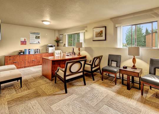 A welcoming leasing office here at Deer Park featuring a wooden desk with guest seating, coordinated chairs, side tables with lamps, and built-in storage cabinets. Warm lighting, neutral wall tones, and large windows create a professional, comfortable space designed for resident support and everyday leasing assistance.
