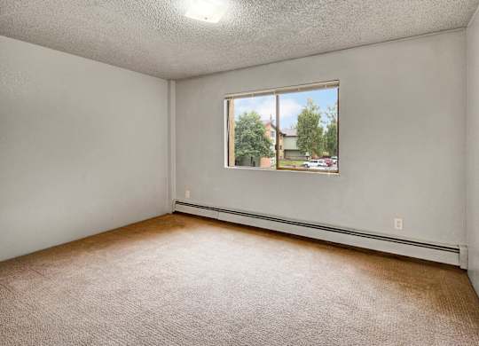 Airy bedroom here at Deer Park featuring wall-to-wall carpet, light neutral walls, and a large window that brings in natural light and outdoor views. A simple layout provides flexible space for bedroom furniture, while baseboard heating and an overhead light add everyday comfort and functionality.