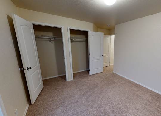 A bright, carpeted bedroom here at Four Seasons featuring soft neutral walls, overhead lighting, and an open layout that offers flexible furniture placement. An open closet with shelving and hanging space provides convenient storage, while nearby doorways connect easily to the rest of the apartment.