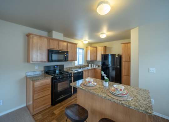 A bright, modern kitchen here at Four Seasons showcasing natural wood cabinetry, granite-style countertops, and a spacious center island with seating, paired with black appliances, warm wood-look flooring, ceiling lighting, and an open layout that offers ample prep space and an inviting flow into the surrounding living area.