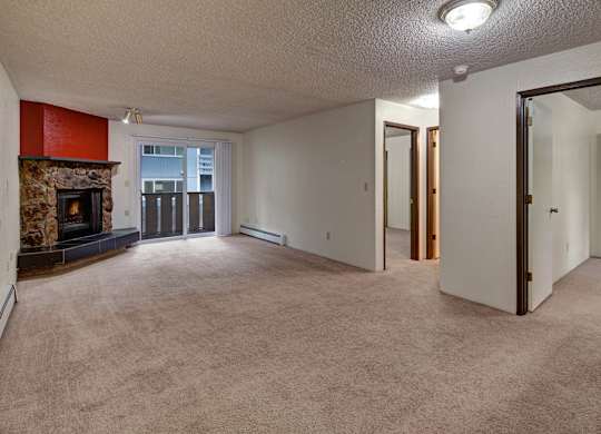 A spacious and inviting living room here at The Greenbriar featuring soft wall-to-wall carpeting, neutral walls, and a cozy stone fireplace with a bold accent surround. The room includes sliding glass doors leading to a private balcony, multiple doorways for easy flow to bedrooms and closets, and overhead lighting that creates a warm, comfortable atmosphere.