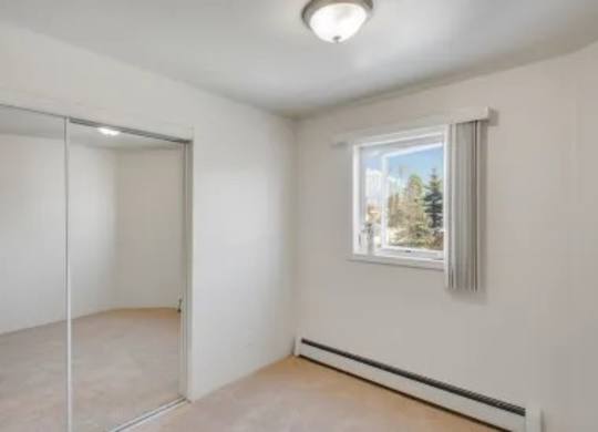 A bedroom here at La Maisonnette with beige carpet and white walls. A window with vertical blinds brings in natural light, and mirrored closet doors provide storage and reflect the space.