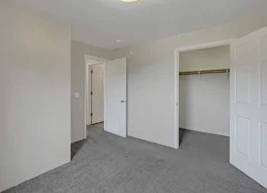A bedroom here at La Maisonnette, with soft carpeting and neutral walls. An open closet with a hanging rod provides storage, and the room connects to a hallway through a standard interior door.