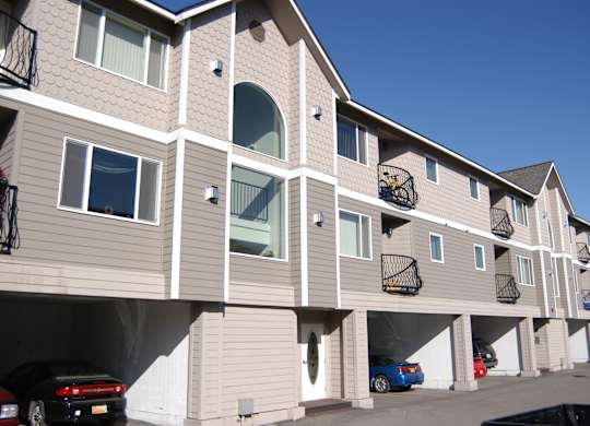 Three-story apartment building here at La Maisonnette with beige siding and white trim. The exterior features multiple private balconies, large windows, and covered parking beneath the building.