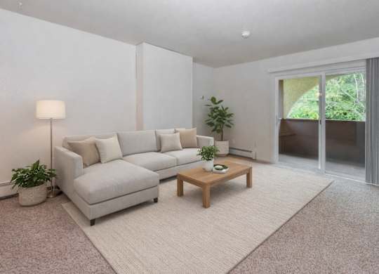 A living room here at Ladera Villa, with beige carpet and white walls. A light-colored sectional sofa sits on an area rug with a wooden coffee table, and a sliding glass door opens to a private balcony with greenery outside.