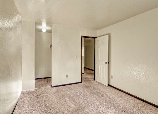 An open and empty bedroom space here at Ladera Villa with beige carpeting and neutral walls. The room features a standard closet space, overhead lighting, and a door leading to the hallway.
