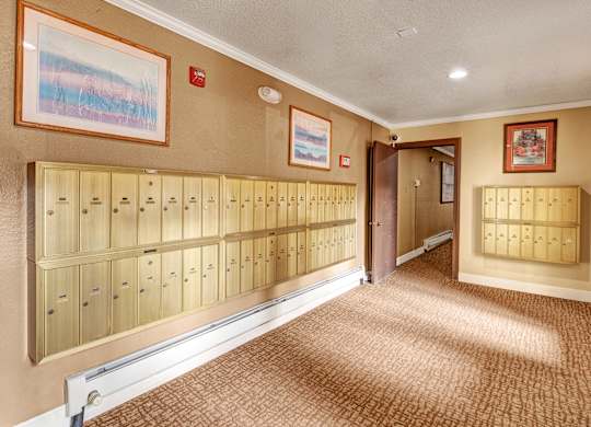 Common area mailroom here at Ladera Villa with beige walls and patterned carpet flooring. Wall-mounted mailboxes line the hallway, with framed artwork and warm overhead lighting creating a welcoming atmosphere.