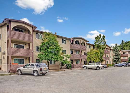 Exterior view here at Ladera Villa featuring a three-story apartment community with beige siding and brown balcony accents. The property includes private balconies, mature trees, and an open parking area, creating a welcoming residential setting.
