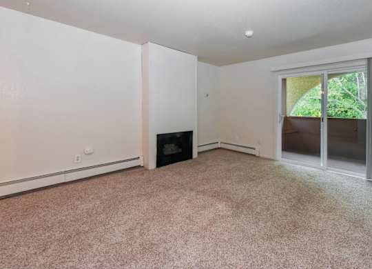 An open-concept and empty living room here at Ladera Villa, with beige carpet and neutral walls. A fireplace is set into the wall, and a sliding glass door opens to a private balcony with greenery outside, creating a welcoming atmosphere.
