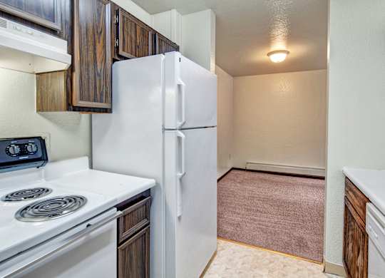 A compact kitchen here at Ladera Villa, with dark wood cabinets, white countertops, and white appliances, including a stove and refrigerator. The kitchen opens to a carpeted dining area with overhead lighting. and soft carpeting.