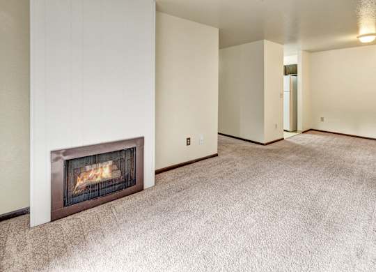 An open and empty living room space here at Ladera Villa, with beige carpet and neutral walls. A built-in fireplace adds warmth, and the open layout leads toward the kitchen and dining area.