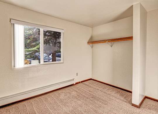 An open and empty bedroom space here at Ladera Villa, with beige carpet and neutral walls. A window provides natural light and views outside, and an open closet with a hanging shelf offers storage space.