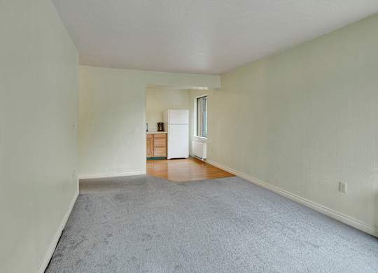 A spacious open living area here at Mendenhall-Tower with soft gray carpeting, light neutral walls, and a connected dining nook featuring warm wood flooring and a bright window for natural light.