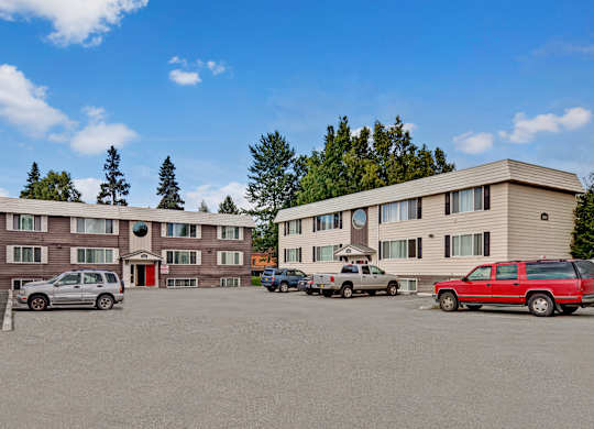 A wide exterior view of Montclair showing multiple three-story apartment buildings with neutral siding, red entry doors, and an open parking lot surrounded by mature trees.