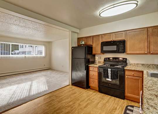 A bright open-concept kitchen here at Montclair with warm wood cabinets, speckled granite-style countertops, and black appliances, overlooking a carpeted living area with a wide window and vertical blinds, creating a cozy and welcoming atmosphere.