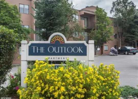 A welcoming entrance sign for Outlook Apartments surrounded by bright yellow flowers, with the apartment building and parking area in the background.