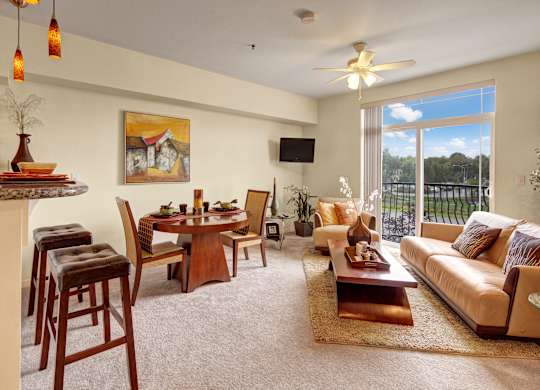 A bright, open-concept living and dining room here at Park Plaza II with beige carpeting, a tan sofa and striped accent chair, and a wooden coffee table on a cozy rug, flowing into a kitchen breakfast bar with pendant lighting.