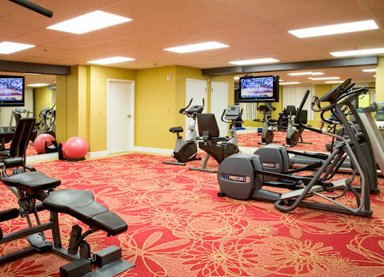 A spacious fitness center with cardio and strength equipment including ellipticals, bikes, and benches, featuring wall-to-wall mirrors, mounted TVs, and bright lighting for an energetic workout environment.