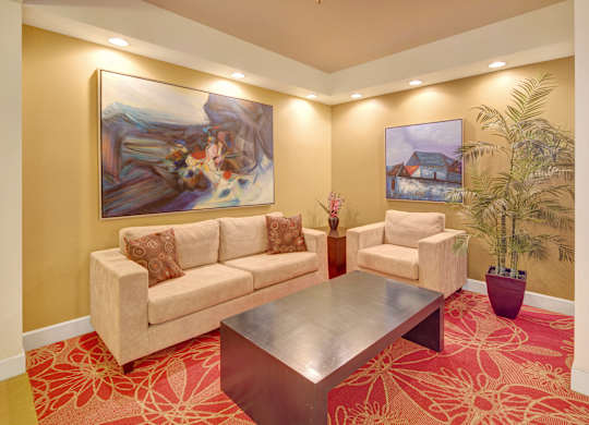 A cozy lounge area here at Park Plaza II with a beige sofa and matching armchair, featuring modern wall art, soft recessed lighting, and a large potted plant for a warm, inviting atmosphere.