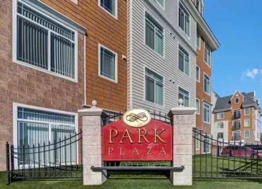 A welcoming exterior sign here at Park Plaza II with the community name displayed in front of the multi-story apartment building, featuring classic siding, large windows, and a clean, well-maintained setting.