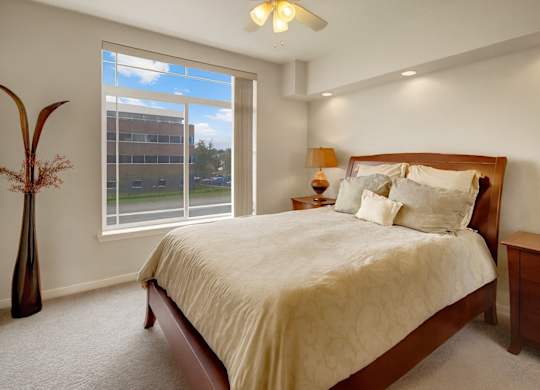 A bright bedroom here at Park Plaza II with plush beige carpeting and a large window with vertical blinds, featuring a cozy bed with neutral bedding, matching wood nightstands, and soft ceiling fan lighting.