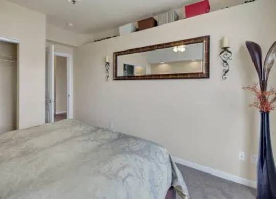 A cozy bedroom here at Park Plaza II with soft neutral carpeting and warm beige walls, featuring a comfortable bed and a large wall mirror that reflects the space for a bright, open feel.