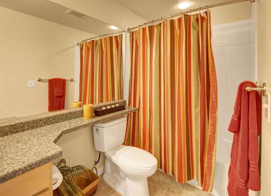 A bright bathroom here at Park Plaza II with a bathtub-shower combo and white tile surround, featuring a wide vanity with speckled granite countertops, a large mirror, and warm striped shower curtains for a cozy, inviting feel.