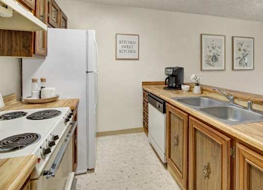 A clean, cozy galley kitchen with warm wood cabinets and butcher-block style countertops, featuring a white refrigerator, white stove, double stainless sink, and simple wall decor with framed botanical prints.