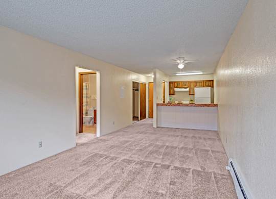 A long, open living and dining area here at Park Plaza with soft beige carpeting and bright white walls, featuring a breakfast bar that overlooks the kitchen with warm wood cabinets, a white refrigerator, and ceiling fan lighting.