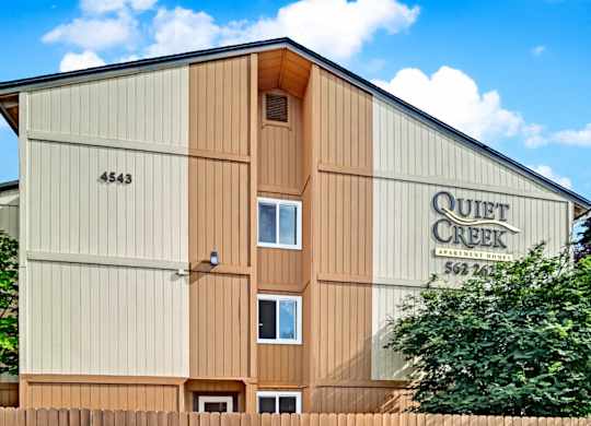 A close-up exterior view here at Quiet Creek featuring the community building with the Quiet Creek Apartment Homes sign, warm neutral siding, and mature greenery under a bright blue sky.