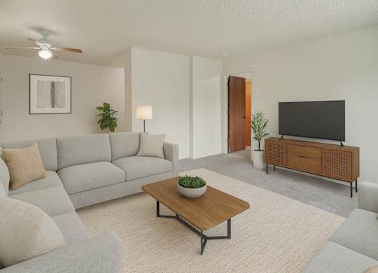 A bright living room here at Rancho Tudor with a light gray sectional sofa, a wooden coffee table, a soft neutral rug, and a modern TV console with simple greenery accents.