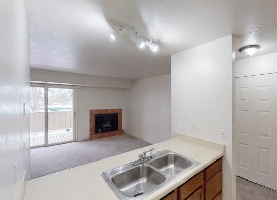 A bright open-concept living space here at Russian Jack with a double kitchen sink and breakfast bar overlooking the carpeted living room. A cozy corner fireplace and sliding glass door lead to a private balcony, creating an airy and inviting feel.