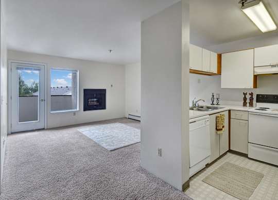 A bright, open-concept living space here at Silver Ridge with soft beige carpet and white walls for a clean, airy feel. A compact kitchen features cream cabinets and white appliances, while the living room includes a cozy fireplace and a glass door leading to a private balcony.