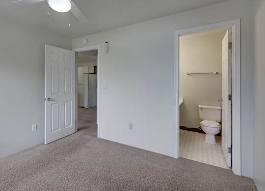 A clean, spacious bedroom here at Silver Ridge with soft beige carpet, light walls, and a bright, simple layout. A ceiling fan with a light adds comfort, while an ensuite bathroom and open doorway leading toward the kitchen create an easy, functional flow.