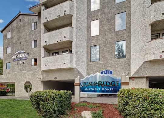 A front exterior view here at Silver Ridge showcasing the community building with private balconies and a clean, modern facade. A Silver Ridge Apartment Homes entrance sign sits in a landscaped area with green grass and shrubs, creating a welcoming first impression.