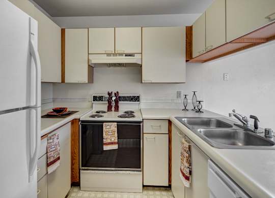 A compact kitchen here at Silver Ridge with cream-colored cabinets, white countertops, and warm wood trim for a cozy, classic feel. A white refrigerator and electric stove sit beneath a range hood, while a double stainless steel sink and dishwasher add everyday convenience.