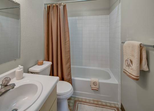 A clean bathroom here at Silver Ridge with a tub-and-shower combo, white tile surround, and a warm neutral shower curtain. A white vanity with a wide countertop sits beside a large mirror, with a toilet and towel bar adding simple everyday comfort.