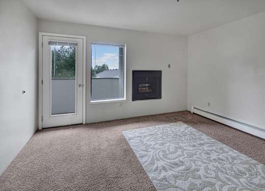 A spacious, empty living room here at Silver Ridge with soft beige carpet and clean white walls for a bright, open feel. A sliding glass door and window bring in natural light, while a built-in fireplace adds a cozy touch and leads out to a private patio or balcony area.