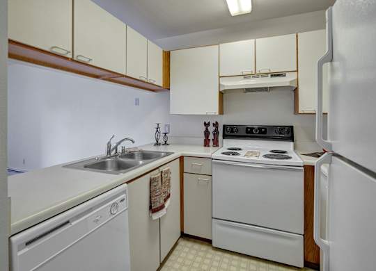 A functional kitchen here at Silver Ridge with cream-colored cabinets, white countertops, and warm wood accents for a classic, inviting look. A white refrigerator, stove, and dishwasher pair with a double stainless steel sink to create a practical and efficient cooking space.