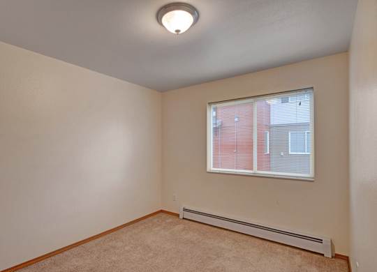 A bright, empty bedroom here at Strawberry Lane with soft beige carpet and warm neutral walls for a clean, cozy feel. A wide window with blinds brings in natural light, creating a simple and comfortable space to make your own.