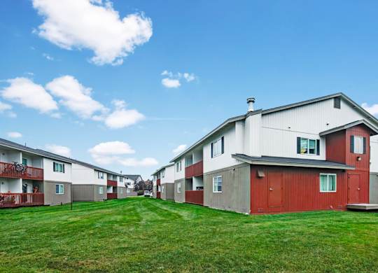 A bright exterior view here at Strawberry Lane featuring multi-level apartment buildings with red and gray siding, wide green lawn space, and a clean, open community layout under a sunny blue sky.