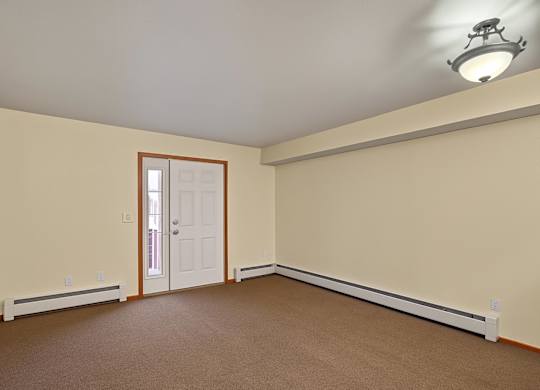 A spacious, empty living room here at Strawberry Lane with soft beige carpet and warm neutral walls for a clean, open feel. A ceiling light brightens the space, while a front entry door with a side window adds natural light and an inviting touch.
