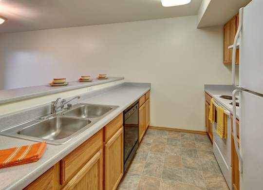 A spacious galley kitchen here at Strawberry Lane with light wood cabinets, neutral countertops, and easy-to-clean tile flooring. A double stainless steel sink, dishwasher, white refrigerator, and stove create a practical layout, with a long breakfast bar offering extra prep space and casual seating.