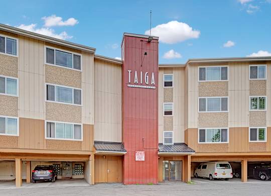 A bold exterior view here at Taiga featuring a multi-story apartment building with a central Taiga Apartment Homes sign, large windows, and covered parking below under a bright blue sky.