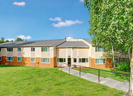 A peaceful exterior view here at Taiga featuring a well-maintained apartment building with large windows, a wide green lawn, and a sidewalk ramp with handrails for easy access under a bright blue sky.