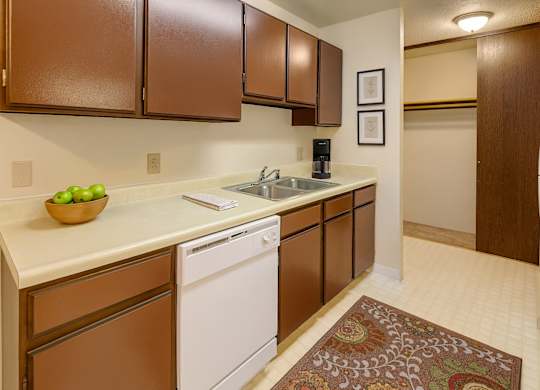 A cozy kitchen here at Taiga with dark brown cabinets, light countertops, a double stainless steel sink, and a white dishwasher, plus an open view into a spacious closet area for added convenience.