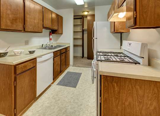 A functional compact kitchen here at Taiga with warm wood cabinets, light countertops, a double sink, and white appliances including a dishwasher, refrigerator, and stove, with extra shelving and closet space nearby.