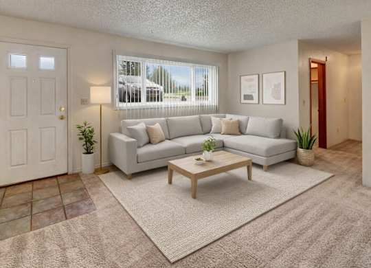 A bright living room here at Terrace on the Lake with a light gray sectional sofa, neutral throw pillows, a wooden coffee table on a beige rug, and a large window with vertical blinds that brings in natural light and outdoor views.