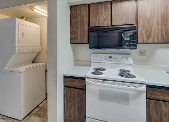 A compact kitchen area featuring dark wood upper and lower cabinets, a white electric stove with coil burners, and a black built-in microwave above it. To the left, a stacked white washer and dryer sit in a recessed laundry nook with overhead lighting, all set against light-colored walls and a tiled floor.